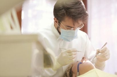A male dentist with a beard and glasses, wearing a white lab coat and blue mask, examines a female patient's teeth.